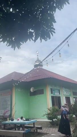 The video shows an exterior view of a mosque with a green wall and a red tiled roof. A silver dome with a crescent moon and star sits atop the mosque. String lights with decorative bulbs are strung across the top of the frame. In the foreground, a person wearing a black hijab is holding a baby. To the left, there is a picnic table with two people sitting at it. A tree with large leaves partially obscures the view of the sky. The overall scene appears to be outdoors during daylight with cloudy skies.