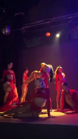 Here is a description of the video:

The video shows a group of dancers on a stage performing a routine. The dancers are all wearing matching red costumes with variations in style. There are 6 women and 2 men in the group. One of the male dancers is holding a woman horizontally above his head. The other male dancer is crouching low on the ground. The women are positioned around them in various poses. The stage is lit with red and purple lights, creating a dramatic atmosphere. There is a disco ball hanging from the ceiling.