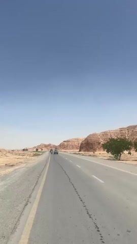 A daytime video shows a long, straight asphalt road cutting through a desert landscape in Kuchlak, Pakistan. The sky is clear and light blue. To the right, barren, reddish-brown rocky hills and mountains stretch into the distance. A single green tree stands out against the arid backdrop on the right side of the road. To the left, the terrain is flatter and sandy.

The road has two lanes with a yellow line marking the left shoulder and white dashed lines separating the lanes. Dark, irregular marks are visible on the road surface, possibly stains. Two vehicles, including what appears to be a white SUV or pickup truck, are seen driving away from the camera in the distance. The overall scene depicts a remote and dry environment under a bright, sunny sky.