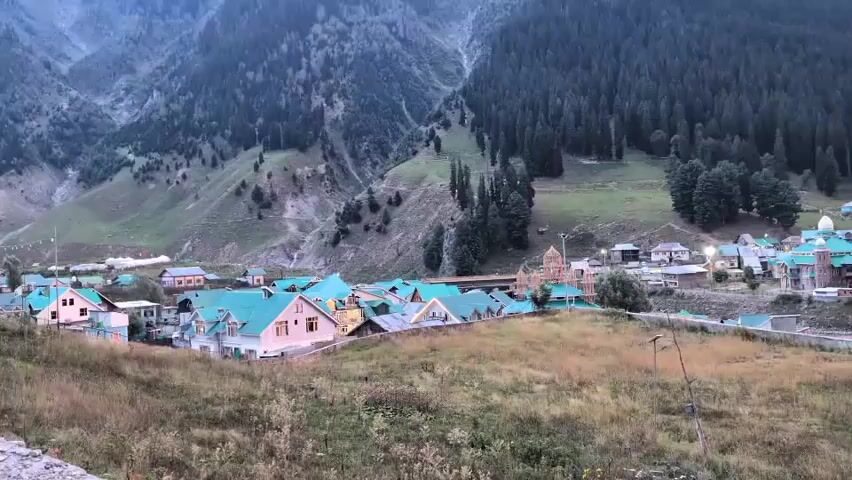 A wide shot captures a village nestled in a valley at the base of a majestic mountain range. The slopes of the mountains are blanketed with a dense forest of pine trees. The village itself consists of numerous buildings with distinctive turquoise roofs, arranged along a central road. Some of the buildings appear to be houses, while others might be lodges or guesthouses, indicated by the presence of various colors and architectural styles. In the foreground, a grassy field with dry vegetation leads towards the village. The sky transitions from a dusky twilight to a dramatic sunset, with vibrant hues of orange and pink illuminating the horizon between the peaks of the mountains. The lighting suggests the video was filmed during either sunrise or sunset, casting long shadows and imbuing the scene with a serene, ethereal atmosphere. There are no people or animals visible in the frame. The overall impression is one of natural beauty and a tranquil rural settlement.