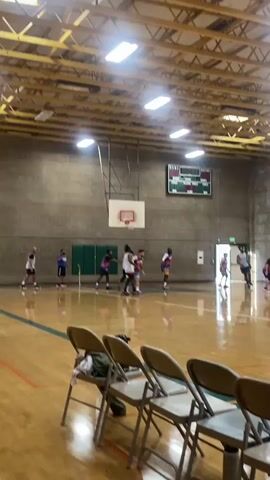 A basketball game is in progress on an indoor court. Several players are on the court, some in uniform, engaging in the game. The court has a polished wooden floor with painted lines. Bleachers with chairs are visible in the foreground. A scoreboard hangs on the wall in the background. The environment appears to be a gymnasium or sports facility.