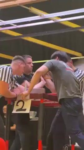 Four men surround an arm wrestling table with a number "2" displayed on the front. Two of the men, wearing striped shirts, are referees or officials. The other two, dressed casually, are arm wrestling. The man in the green shirt and black pants appears to be winning. They are indoors with a black and yellow ceiling structure visible above.
