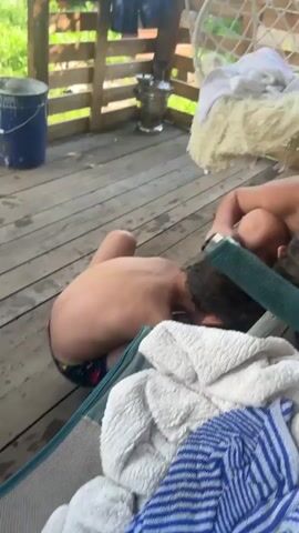 The video shows a close-up of a person's leg with several red bumps, possibly insect bites. The person is wearing a light-colored clog. In the background, wooden planks of a deck are visible, along with a person wearing patterned shorts and part of a blue chair with a towel. The overall environment appears to be outdoors. The action involves the camera zooming in on the leg and the red bumps.