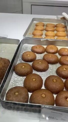 A pan of golden-brown pastries, some cut into triangular pieces, sits on a silver tray. Behind it, other pans hold rows of similar pastries, some sprinkled with green pistachios. The pastries are arranged in a display case, with a white countertop visible in the background.