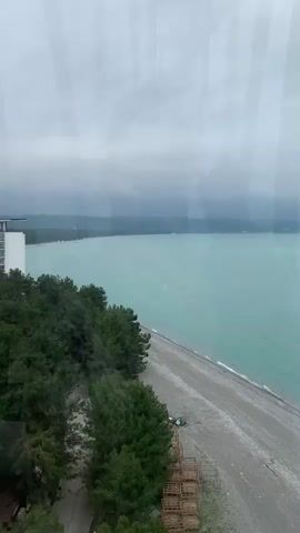 The video shows a view of the coastline from a high vantage point, likely from a window or observation deck. The sky is overcast and the water is a milky turquoise color. A beach runs along the coastline, and there are trees and some man-made structures visible. The camera pans slightly, revealing the frame of the window. The video is still, with no people or significant actions observed.