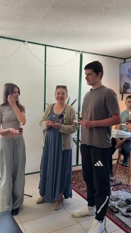 A group of people are gathered indoors, each holding a small cupcake with a lit candle on top. The individuals are of various ages, including an older woman in a blue dress, a young man in a grey t-shirt and track pants, and a young woman in a grey cropped top and sweatpants. Another woman is speaking into a microphone, wearing a beige shirt and teal wide-leg pants. The room appears to be a studio or indoor event space, with a rug on the floor, a TV screen displaying text, and music equipment such as a guitar and speakers in the background. People are smiling and looking at each other, suggesting a celebratory mood. A small dog is standing near the woman with the microphone.