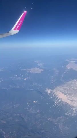 The video shows a view from an airplane window as it flies over a mountainous, coastal region. The airplane's wing, with a pink Wizz Air logo on its tip, is visible in the foreground, extending from the left side of the frame. Below the wing, the earth unfolds with rugged mountain ranges covered in vegetation and dotted with scattered settlements. A body of water, likely the sea, meets the coastline, and further inland, a lake or reservoir is visible. The sky is a clear, vibrant blue, indicating a sunny day. The perspective suggests the plane is descending or in a level flight path, with the ground appearing to recede into the distance. There are no people or significant interactions depicted, only the vast landscape seen from a high altitude.