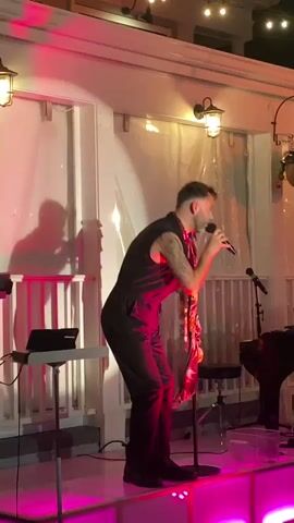 A man in a black jumpsuit, with a scarf draped around his neck, stands on a stage with pink lighting. He is holding a microphone and appears to be singing or performing. To his left, a keyboard and a laptop are visible on a stand. In the background, there are string lights and a white wall with windows. The man's shadow is cast on the wall behind him. He is holding the scarf with his left hand and the microphone with his right. The overall scene suggests a live musical performance in an indoor or covered outdoor venue.