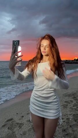 A young woman with long red hair takes a selfie on a beach at sunset. She holds a smartphone with a light attached to it. She is wearing a white, see-through dress with long sleeves. The sky is a mix of dark clouds and vibrant colors from the setting sun. The ocean waves are gently lapping against the shore.
