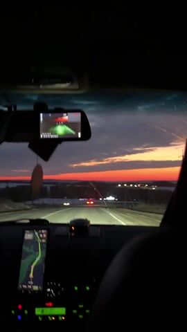A car drives down a road as the sun sets. The sunset is a beautiful mix of orange, pink, and purple hues. The car has a dashcam and GPS system running. The GPS shows a route on a map. The road appears to be well-maintained and there are other cars in the distance. The driver's seat is visible, but no driver is seen. The environment surrounding the road is flat with low lying buildings.
