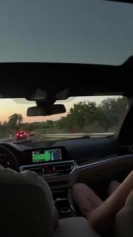 The video shows the interior of a car driving on a road. The perspective is from the back seat, looking forward. The sky is visible through the sunroof, with a sunset casting a warm glow. Trees and vegetation line the road, and other cars can be seen ahead. The car's dashboard is visible, including a navigation screen displaying a map. The interior lights of the car are on, with subtle ambient lighting. Two individuals are partially visible in the front and back seats. One person in the front passenger seat has their legs extended, and another person in the back seat is seen from the side with their head turned away from the camera. The overall atmosphere suggests a scenic drive during dusk.