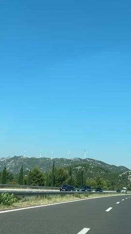 The video shows a view from a car traveling on a highway. The highway has two lanes visible, with white lines marking the lanes. There is a metal guardrail separating the highway from the scenery. In the background, there is a green, hilly landscape dotted with trees. On top of the hills, several wind turbines are visible, spinning slowly. The sky is a clear, bright blue. Several cars are driving on the highway in the same direction as the camera. Some cars are black SUVs, one is a white van, and another is a white truck. The time stamp indicates the video is from the very beginning of the day.