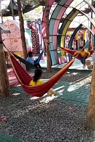 The video shows an outdoor recreational area with several hammocks suspended between trees. In the foreground, a red hammock contains two people, one in a yellow shirt and another in a blue shirt. To the right, another hammock is occupied by a person in a red shirt. In the background, a colorful, arched structure resembling a rainbow is visible, along with other people and structures. The ground is covered in gravel and patterned paving stones. The scene appears to be a playground or park designed for leisure activities.