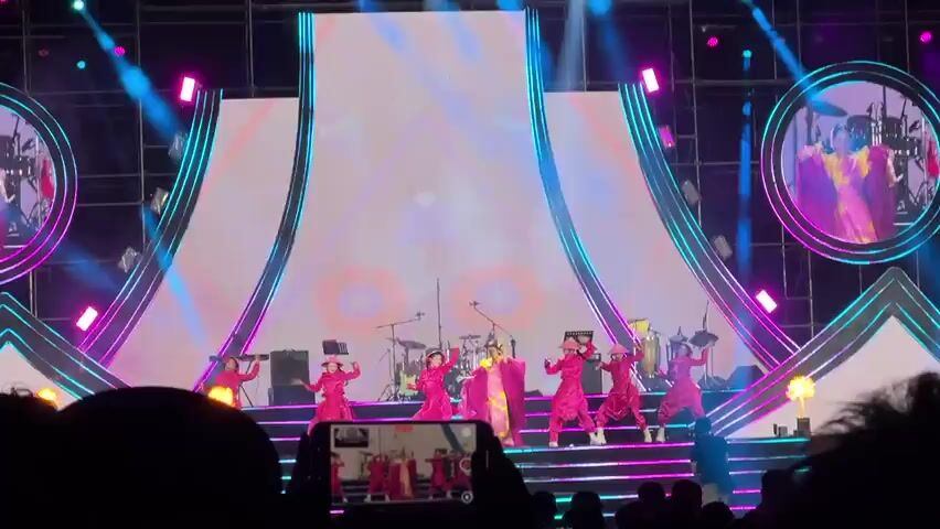 A vibrant live performance unfolds on a stage, featuring a main female performer and six accompanying dancers. The main performer, positioned centrally, wears an elaborate yellow garment beneath a flowing, bright pink or magenta cape. She appears to be singing or leading the performance. The six dancers, who appear to be children or young adults, are dressed in matching hot pink outfits and wear traditional Vietnamese conical hats (nón lá). They perform synchronized choreography around the main artist.

The stage is brightly lit with various colored spotlights, primarily in pink, purple, and blue hues. A large LED screen serves as the backdrop, displaying dynamic, abstract graphics including stylized red and blue floral patterns, geometric shapes, and neon lines that change throughout the performance. Behind the performers, a band set-up is visible, including a drum kit, keyboards, and percussion instruments.

The atmosphere is energetic and festive. A section of the audience is visible at the bottom of the frame, with one person actively recording the performance on a smartphone, indicating an audience-attended event. The performance is taking place in Măng Đen, Vietnam, suggested by the provided context, which aligns with the cultural attire worn by the dancers.