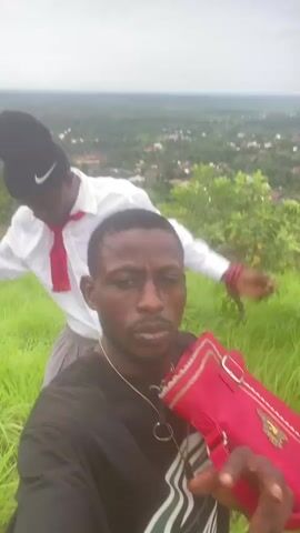 A selfie video shows two young black men outdoors on a grassy hillside with a village visible in the distance. The man in the foreground, wearing a black t-shirt with a white graphic design and a gold chain, is holding a red purse and smiling at the camera. Behind him, another man wearing a white dress shirt, a red tie, and a black beanie with a white Nike logo adjusts his tie. The sky is overcast.