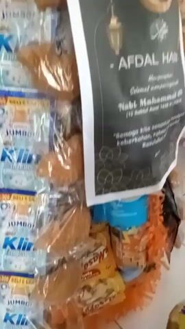 The video shows a display of food items wrapped in plastic and arranged in a decorative manner. The display appears to be in a market or store setting, with other food products and shelves visible in the background. The central item is a tiered arrangement of what look like fried dough balls or small pastries, wrapped in plastic, and adorned with large green leaves. One of the items in the display is a packet with Islamic calligraphy and a crescent moon symbol, suggesting it might be related to a religious occasion. The lighting suggests an indoor environment, possibly a small shop or a market stall. There are no people clearly visible interacting with the display in the foreground, but the background suggests a public space.