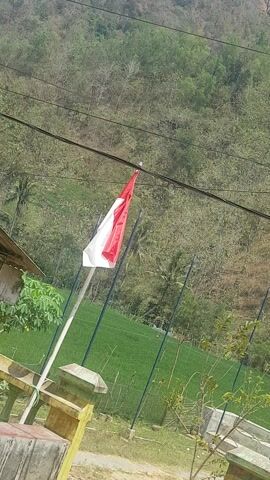 A red and white flag, likely the flag of Indonesia, is flapping in the wind on a flagpole. The flag has two horizontal bands, red on top and white on the bottom. The flagpole is white and extends from the bottom of the frame upwards towards the flag. Behind the flag, there is a lush green field, possibly a sports field or park, bordered by dark poles. Further in the background, a dense forest covers a hillside, with trees appearing to be in various stages of leaf cover. Power lines are visible running horizontally across the upper left portion of the frame. In the lower right corner, a section of a building or structure is visible, constructed with yellow and grey materials, suggesting it might be part of a park or recreational facility. The scene appears to be set outdoors during the daytime, with bright sunlight illuminating the landscape. The overall impression is one of a tranquil, natural setting with a touch of national pride represented by the flag. The location context places this scene in Tinatar, Indonesia.