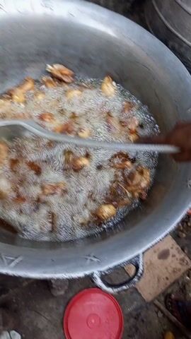 A close-up, medium-angle shot depicts pieces of food, possibly chicken, being deep-fried in a large, silver-colored pot or wok. The pot is filled with bubbling oil, and a silver, slotted spoon is partially submerged in the oil, used to stir or monitor the cooking process. The food pieces appear to be browning and are scattered throughout the oil. The pot has the inscription "FRANCIS *" on its side. The setting appears to be outdoors, with a red container visible near the bottom of the frame.