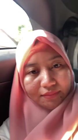 A close-up selfie video shows a young woman wearing a pink hijab and a white shirt. She is sitting in the passenger seat of a car, as indicated by the car's interior visible in the background. The lighting suggests it is daytime, with sunlight illuminating her face. The woman is looking at the camera and smiling. The video is brief, lasting only a few seconds.