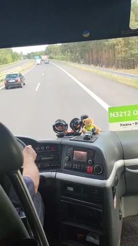 The video is filmed from the passenger's perspective inside a bus, looking forward. The bus is traveling on a highway. The dashboard of the bus is visible, with a steering wheel, various buttons and knobs, a radio, and a screen. A driver's hands are visible on the steering wheel. On the dashboard, there are two small fans, a teddy bear, and a small toy car. A Ukrainian flag sticker is visible at the top of the windshield. A green sign with white text is visible on the right side of the dashboard, partially obscured, but the text "N3212 ОД" is legible. Outside the bus, the highway is visible with other vehicles, including cars and trucks, and a line of trees along the side of the road. The sky appears overcast. The video captures the mundane activity of bus travel.