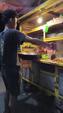 A young man with dark curly hair stands on the left side of the frame, wearing a dark t-shirt and shorts. He is facing a yellow food cart and is in the process of cooking. He is holding a spatula or stir-fry tool in his right hand and is stirring food in a large, dark wok that is positioned over a burner on the cart. The food cart has shelves and compartments, with various containers and utensils visible. A bright light bulb illuminates the cooking area from above. The background is dark, suggesting it is nighttime. The overall scene depicts street food preparation.