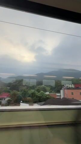 The video captures a view from a moving train window. The scene outside consists of rolling green hills and trees, with a cloudy sky overhead. There are some buildings visible, including a brick building with a sign in what appears to be Korean. In the foreground, the reflection of the train's interior, including lights and seats, is visible on the window, creating a slightly blurred effect as the train moves. The perspective is from a seated position within the train, looking outwards.