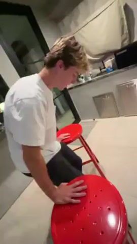 A fair-skinned person with short blond hair, wearing a white t-shirt and dark pants, is positioned with their hands on the seat of a red stool, attempting a push-up. The stool has a round seat with multiple holes. The person is inside a room with a tiled floor and a counter in the background. A large window is visible, reflecting an outdoor scene. The camera is held low, focusing on the person performing the exercise.
