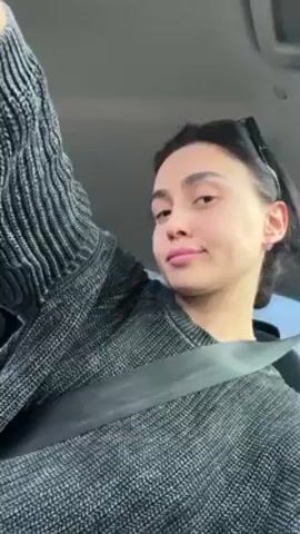 A woman is in the driver's seat of a car, wearing a gray sweater and a seatbelt. She has dark hair pulled back and is looking at the camera while lip-syncing to music. Her right arm is raised, revealing a tattoo on her forearm. The car interior is visible, including the steering wheel and dashboard. The sky is visible through the windshield.