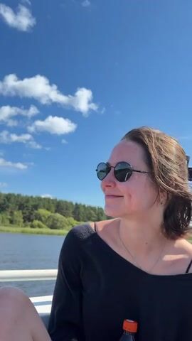 A medium shot shows a woman with short brown hair wearing sunglasses and a black shirt. She is looking to the left of the frame with a slight smile. The background is a bright blue sky with white clouds, and a body of water with trees on the other side.