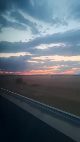 The video shows a view from a moving vehicle, likely a train or car, during sunset. The sky is filled with dramatic clouds, some dark and others tinged with orange and pink from the setting sun. Below the sky is a vast, open landscape, appearing to be fields or plains. There are a few scattered trees and some distant hills or mountains. A prominent feature is the edge of the vehicle's window or frame, which runs along the bottom of the screen, and a billboard or sign is visible passing by in the distance. The overall impression is one of travel and observing a natural landscape during the twilight hours.