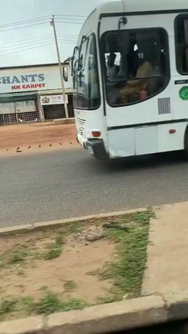 The video captures a street scene, likely in a tropical or subtropical region, characterized by dusty reddish-brown earth and sparse green vegetation along the roadside. A white bus moves from left to right across the frame. In the background, buildings with corrugated metal roofs and what appear to be businesses or workshops are visible. Several people are seen walking or standing near these buildings. There are also motorcycles and what look like three-wheeled utility vehicles parked along the roadside. The sky is overcast. The camera appears to be mounted inside another vehicle, possibly the one following the bus, as indicated by the blurred foreground elements and reflections on a glass surface. The overall impression is of a bustling, everyday street scene in a developing area.