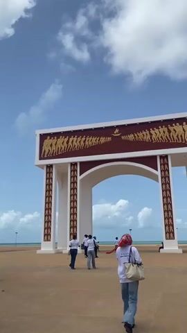 The video shows a grand, white-and-burgundy archway with golden reliefs depicting a procession of figures and a ship, set against a bright blue sky with scattered clouds. People walk around the archway, and in one instance, a person carries a blue basket of goods on their head. The setting appears to be a beachside area with the ocean visible in the background. There is no audio.