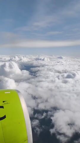 The video is a view from an airplane window. The camera is pointed downwards and slightly forward. The foreground is dominated by the bright green engine cowling of the airplane, which takes up the left side of the frame. Beyond the engine, a vast expanse of white, fluffy clouds stretches out to the horizon. The sky above is a clear blue with thin, wispy white clouds scattered across it. The sun appears to be shining from the right, casting shadows on the clouds. The overall impression is one of flying high above the earth on a clear day.
