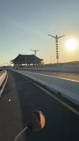 The video shows a view from the perspective of someone riding a motorcycle. In the foreground, a motorcycle's side mirror reflects the rider and the bright, setting sun. The road ahead is asphalt with yellow and white lane markings. Along the right side of the road, a concrete barrier separates the lane from what appears to be a toll plaza or entrance to a bridge. The toll plaza has a distinctive, traditional roof structure resembling a pagoda. The sun is low in the sky, casting a warm glow and long shadows. Streetlights with decorative, ornate tops are visible on both sides of the road. The overall environment appears to be an outdoor road setting, possibly in a location with architectural influences.