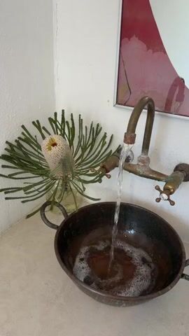 The scene presents a small, white-walled corner of a room with a sink arrangement. The countertop is light-colored. A tarnished, rounded, dark brown, shallow metal bowl with handles serves as the sink.  Water pours from an antique-looking brass faucet, filling the bowl with bubbly liquid.  A glass jar containing an unusual, spiky-leafed flower with a white and yellow center sits to the left of the sink. Above the sink hangs a framed piece of artwork with red and white hues.  The room appears well-lit, focusing attention on the sink area and its unusual design.