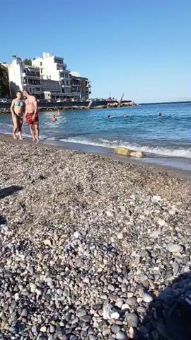 The video captures a beach scene. In the foreground, a collection of pebbles and some beach items like a towel and a bag are visible. The middle ground shows a couple walking on the pebbled shore, with the woman in a bikini and the man in swimming trunks. The background reveals a lively beach with people sunbathing on lounge chairs, swimming in the ocean, and children playing in the water. Buildings with a Mediterranean architectural style are perched on a hillside overlooking the beach. The sky is clear and blue, indicating a sunny day.