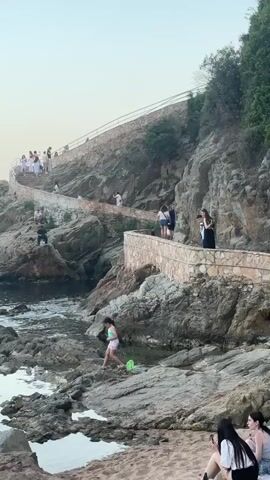The video shows a rocky coastline with a paved walkway winding up a hill. Several people are visible on the walkway and on the rocks near the water. One person, a child, is wading in the shallow water near the shore with a green bucket. Other people are walking along the path, some are taking pictures, and a group is sitting on the beach in the foreground. The scene appears to be a public space, possibly a park or a scenic viewpoint. The weather seems to be mild and sunny.