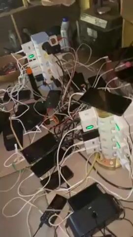 The video shows a large number of cell phones and power banks charging on a surface. Many cords are visible, connecting phones to power sources. The scene is cluttered. Some of the phones have flashing LED lights. There are also multiple charging tower power strips. The floor and surface are covered with the devices and cables.