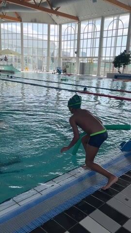 A child with a swim cap jumps into an indoor swimming pool holding a green pool noodle. Another child with a yellow pool noodle swims nearby. The indoor pool has large windows and several other people swimming in it. The pool has lane dividers. There is a lifeguard stand in the background and a staircase.