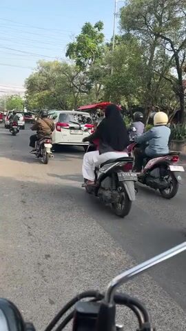 A street scene with several motorcycles and a car on a paved road. The vehicles are moving in the same direction. A person wearing a hijab is riding a motorcycle in the foreground. There are trees and buildings visible on the side of the road. A food cart is set up under a red canopy. The sky is clear and blue.