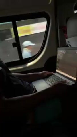 A person sitting in a van is working on a laptop. The person is wearing a black t-shirt. The laptop screen displays lines of code. Outside the van window, scenery goes by.