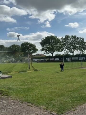 A video shows a wide shot of a green grassy field with a soccer goal on the left and a smaller goal on the right. A child with curly blonde hair wearing a dark blue jacket and light blue pants stands near the smaller goal on the right, looking at it. In the background, beyond the field, there are buildings and trees. Several people are visible in the background, some sitting at tables, others standing. A light pole with multiple lights is seen behind the left soccer goal. The sky is partly cloudy with patches of blue. The video slowly pans to the left.