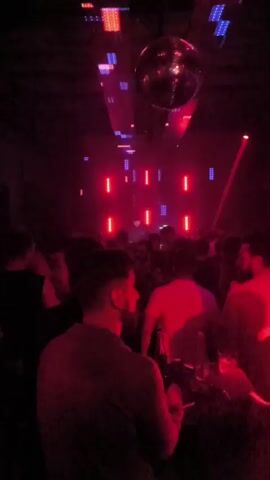 The video displays a dimly lit nightclub with vibrant red and purple lights. A disco ball hangs from the ceiling, reflecting the lights. Several people are visible, with their backs mostly turned to the camera, suggesting they are facing a stage or DJ booth. Some individuals appear to be standing, while others are seated at tables, with drinks in front of them. The overall atmosphere suggests a lively party or clubbing scene, with music implied by the flashing lights and the presence of a DJ setup.