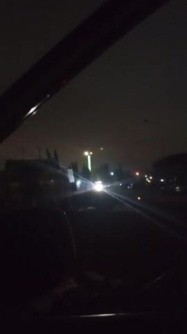 The video shows a nighttime view from inside a vehicle, looking out through the passenger window. The sky is dark, and the road ahead is illuminated by streetlights and the headlights of approaching vehicles. There are buildings and trees visible on the sides of the road, as well as several cars and trucks. The perspective suggests the vehicle is moving, as indicated by the slight motion blur and the way the lights stretch into streaks. The overall atmosphere is dark and urban.