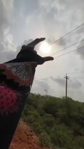 The video begins with a person's hand outstretched towards the sun, partially obscured by clouds. The hand, adorned with a black sleeve featuring a graphic design in white, red, and yellow, reaches toward the bright sun above a landscape of green vegetation and telephone poles. As the camera pans out, the hand remains in the foreground, revealing a dusty, orange-red path adjacent to a paved road. Vehicles, including a white car, travel on the paved road. The sky is overcast with a mix of gray clouds and patches of sunlight, setting a diffused light across the scene. The environment appears to be a rural or suburban area.
