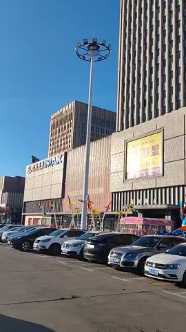 A daytime outdoor shot shows a tall building with a modern facade. The building appears to be a commercial establishment, possibly a shopping mall or office building, as indicated by signage. In front of the building, there is a row of parked cars. The cars are of various makes and models, suggesting a public parking area. Some of the cars have what appear to be delivery boxes or bags in or on them, hinting at potential delivery services being active in the area. Beyond the cars, there are some colorful decorations and possibly amusement rides, suggesting a festive or event atmosphere. A tall street light stands prominently to the right of the frame. The sky is clear and blue. The overall impression is of a busy urban environment.