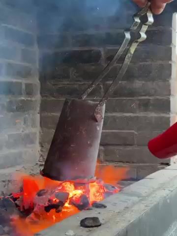 The video shows a person holding a metal cylinder with tongs over a fire in a brick structure. Another person uses a red and black hairdryer to blow air into the fire. The fire is burning intensely, and smoke is rising from it. The scene is outdoors, and the background shows a blurred view of a building and trees.