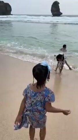 The scene is a beach on an overcast day. Two young girls are seen playing on the sandy shore, running toward the water's edge. One is wearing a blue floral dress and a blue bow in her hair, the other has on a black and white polka dot dress. They interact with two adults, a man and a woman, who are sitting in the shallow water. The water is clear and calm with gentle waves. A large, rocky formation rises from the sea in the distance. The beach appears clean and well-maintained.