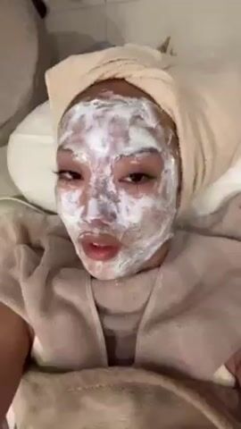 A woman with a white cream face mask and a towel wrapped around her head is lying down and speaking to the camera. She's wearing a robe or towel draped over her body. The mask covers her entire face. She has eyelashes on. She is speaking, stating that she came to a clinic to do a facial treatment. The background shows a pillow and possibly a chair. The lighting is bright.