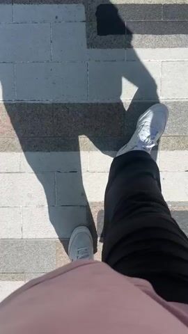 A person is walking along a pedestrian street, capturing a first-person view of their feet and the ground before panning up to reveal the full scene. The ground is paved with a striking design resembling a long set of piano keys, stretching down the center of the street. The person's shadows, along with other shadows, are cast prominently on the ground, indicating a sunny day.

The street is lined with modern buildings, some with large windows and vibrant colored facades (e.g., turquoise and dark green). There are numerous people walking in both directions, and some are seated on benches along the sides of the street. Trees are planted intermittently. In the distance, large mountains are visible under a clear, blue sky, suggesting a mountainous location. One building has the word "PERFUMERIES" written vertically on its side. The overall atmosphere is that of a bustling urban center, possibly a shopping district.